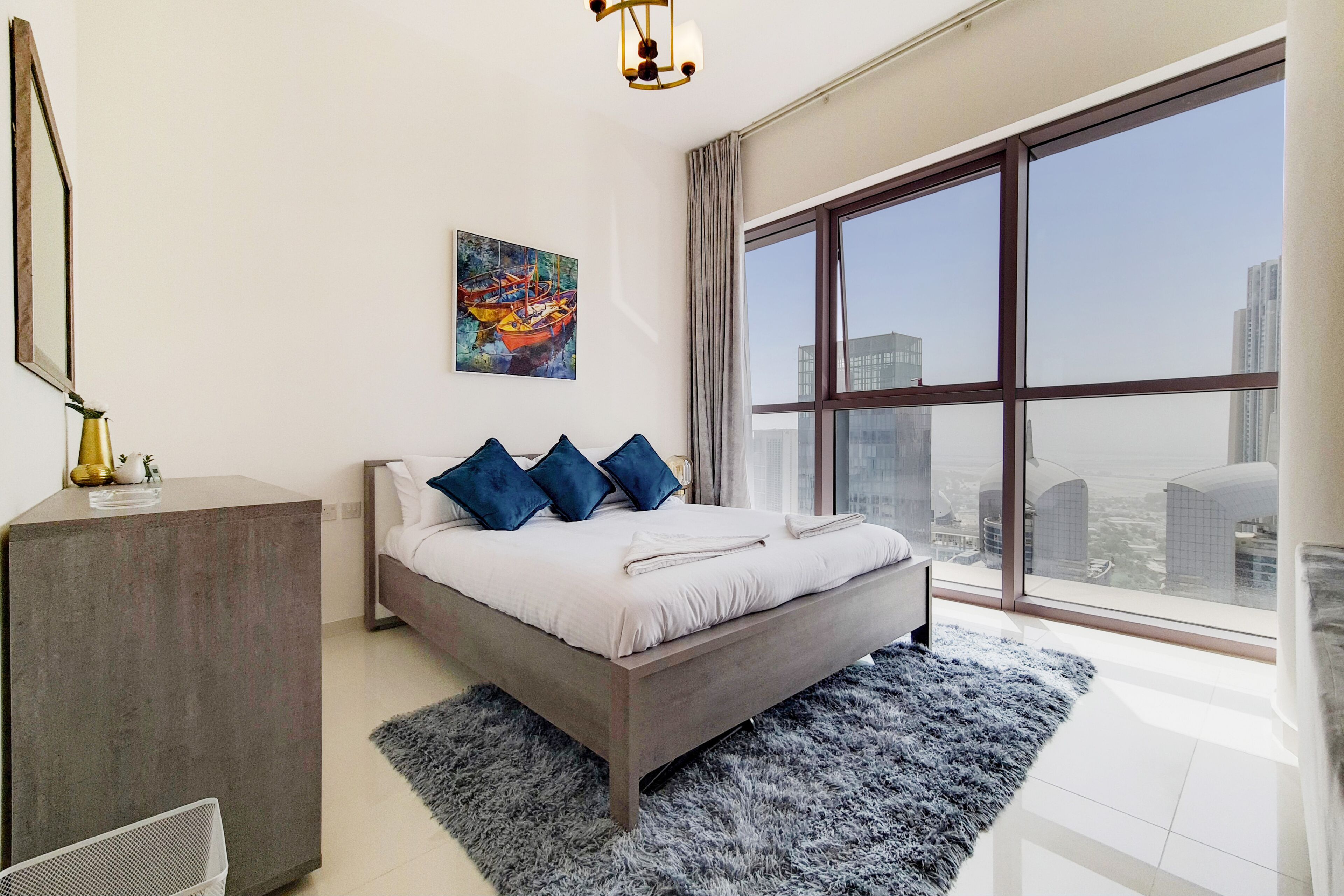 A modern bedroom with a large bed, a grey rug, a painting, and a view of the city through a large window.