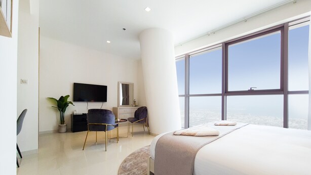 A modern hotel room with a large bed, a desk with a chair, a TV, and a large window offering a city view. - GreenFuture - Luxe Studio In Trade Center With Downtown Views (Dubai)