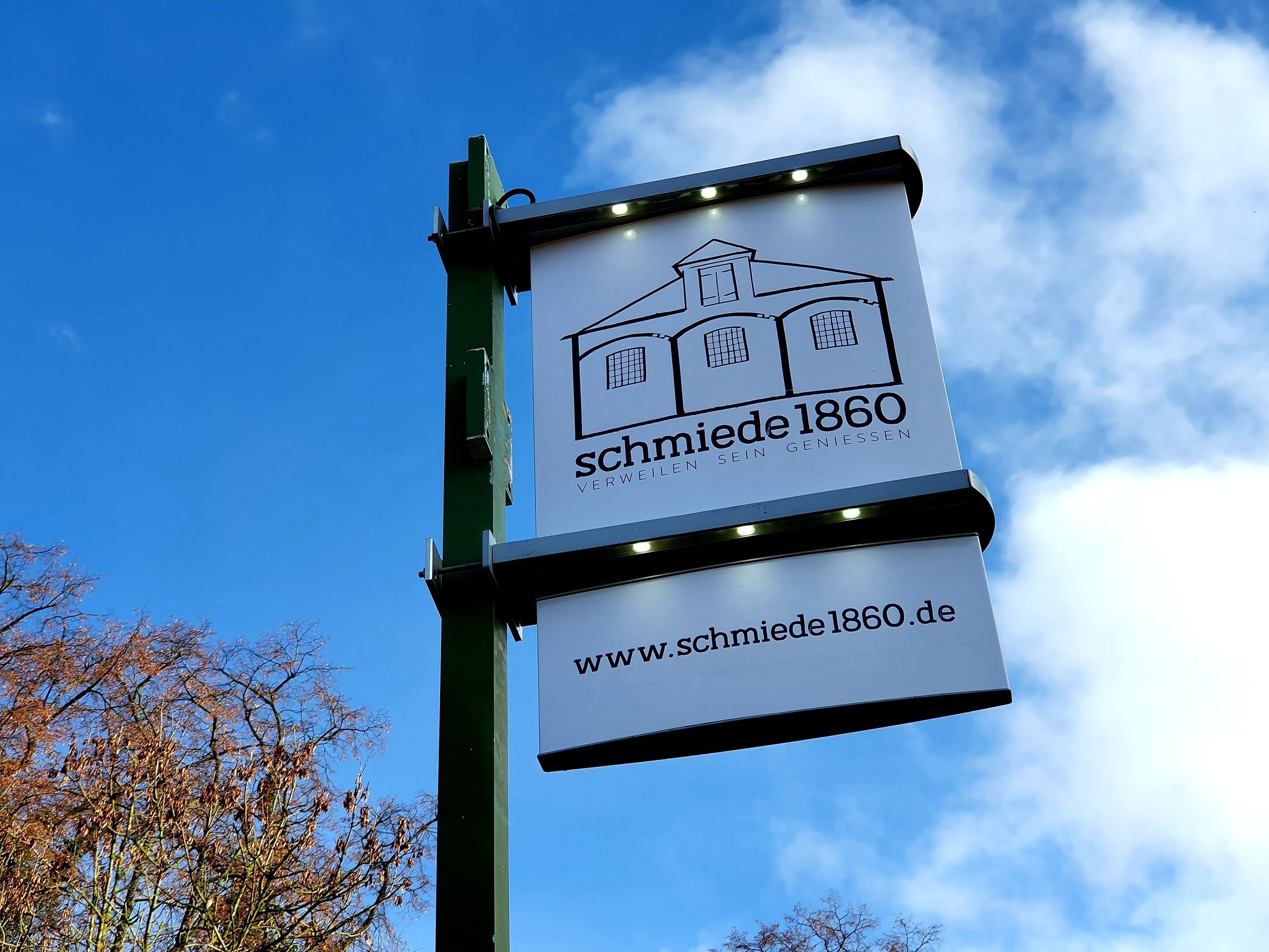 Et skilt med en illustration af en bygning og teksten 'schmiede 1860' samt en hjemmesideadresse.