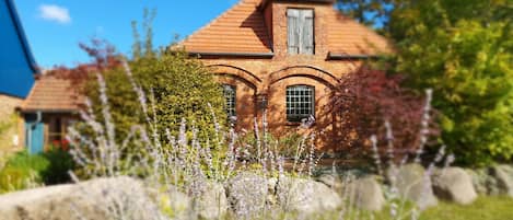 Une maison en briques avec un toit à deux versants et des fenêtres en arc de cercle, entourée d’une végétation luxuriante et de plantes à fleurs.