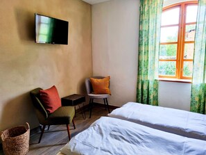 Quarto de hotel com cama, uma cadeira, uma mesinha, televisão fixada na parede e uma janela com cortinas.