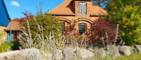 A brick house with a pitched roof and arched windows, surrounded by lush greenery and flowering plants.