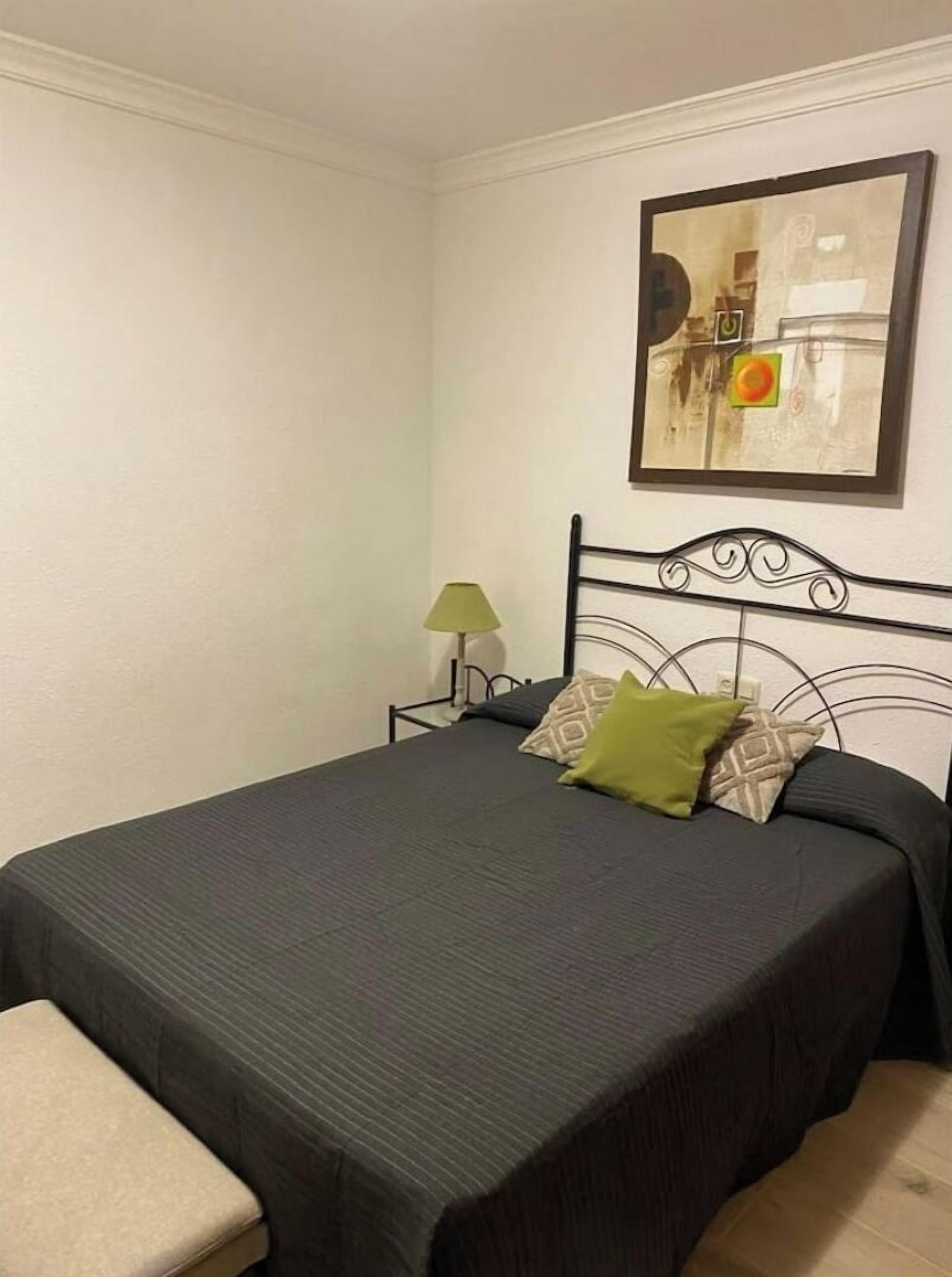 A neatly made bed with a dark bedspread and two pillows, a small bedside table with a lamp, and a framed artwork on the wall.