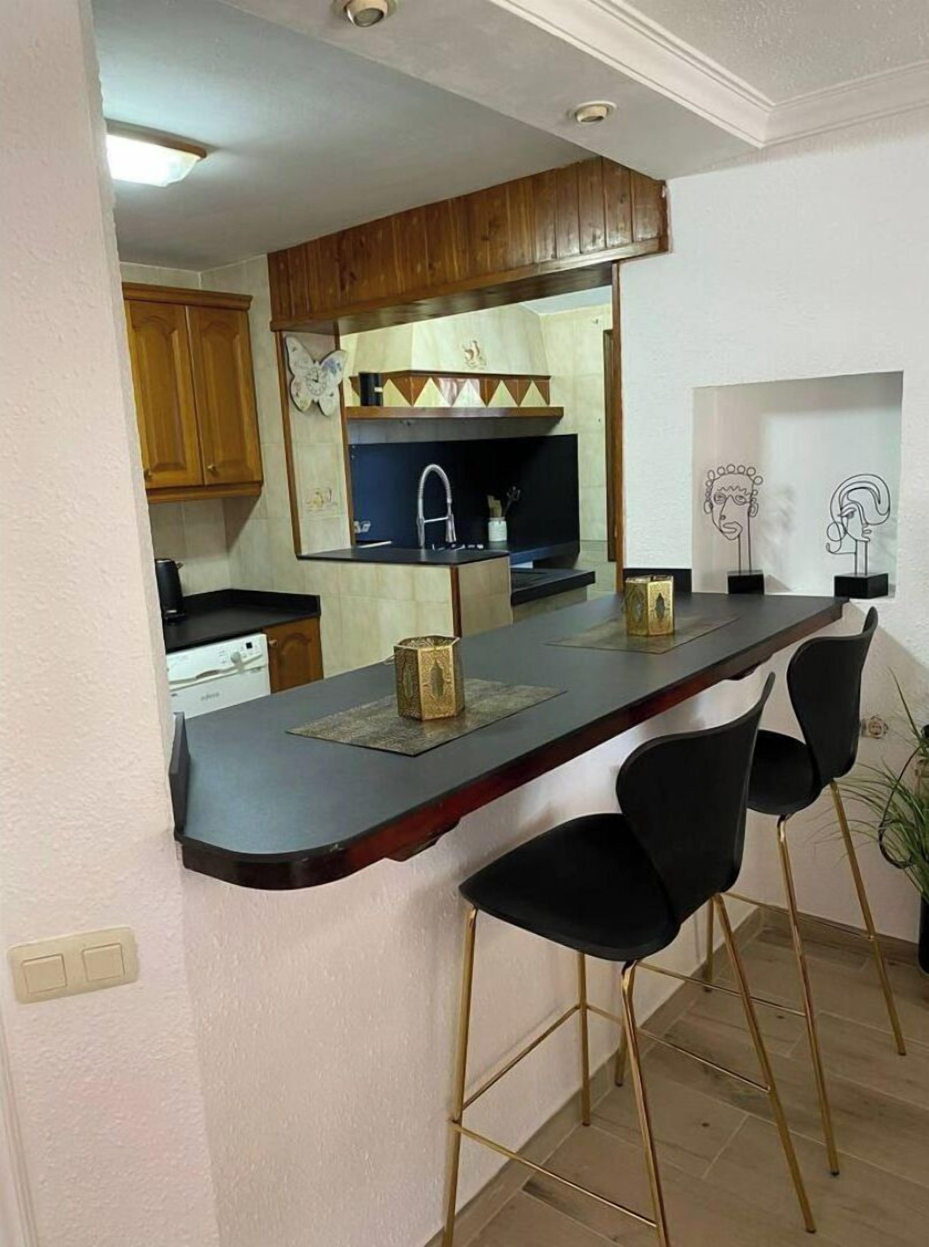 Une cuisine compacte avec des meubles en bois, un plan de travail noir et un évier encastré. On y trouve deux tabourets de bar noirs et un élément décoratif mural.