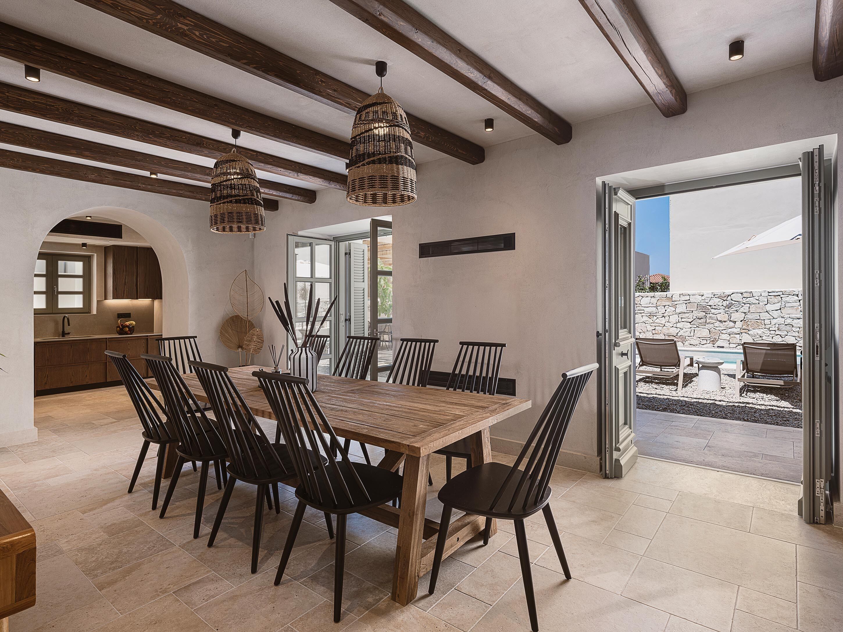 Uma sala de jantar ampla, com mesa de madeira e cadeiras pretas, uma cozinha com janela e vista para uma piscina através de uma porta aberta.