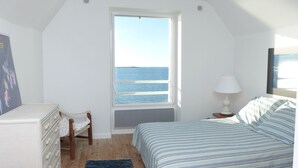 Ein Schlafzimmer mit großem Fenster zum Meerblick, einem Bett mit gestreifter Bettwäsche, einem Nachttisch mit Lampe und einem Sessel mit Kissen.