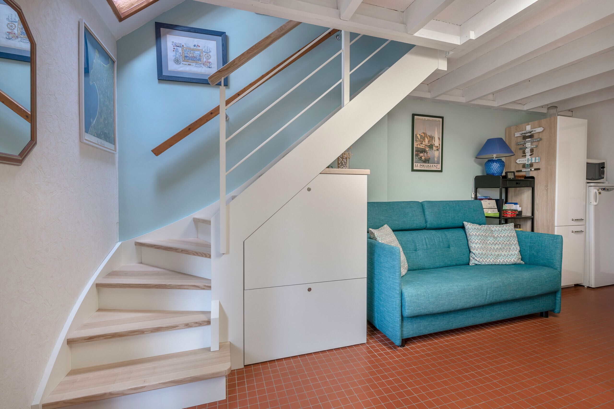 Una escalera con escalones de madera y paredes blancas, un sofá azul aguamarina con almohadas y un refrigerador.