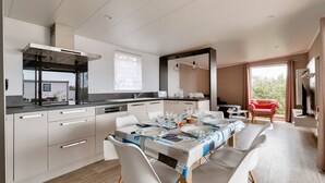 Una cocina moderna con muebles blancos, una zona de comedor con una mesa dispuesta para cuatro personas, y una silla roja junto a la ventana.