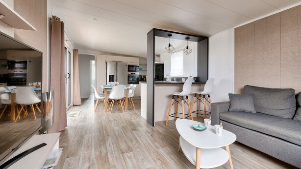 Una sala de estar moderna con un sofá gris, una mesa de comedor blanca con sillas y una cocina al fondo.
