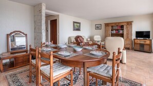Un menjador amb una taula de fusta preparada per a quatre persones, envoltat de cadires amb seients amb coixí. Es veu una columna de pedra i una biblioteca plena de llibres al fons.