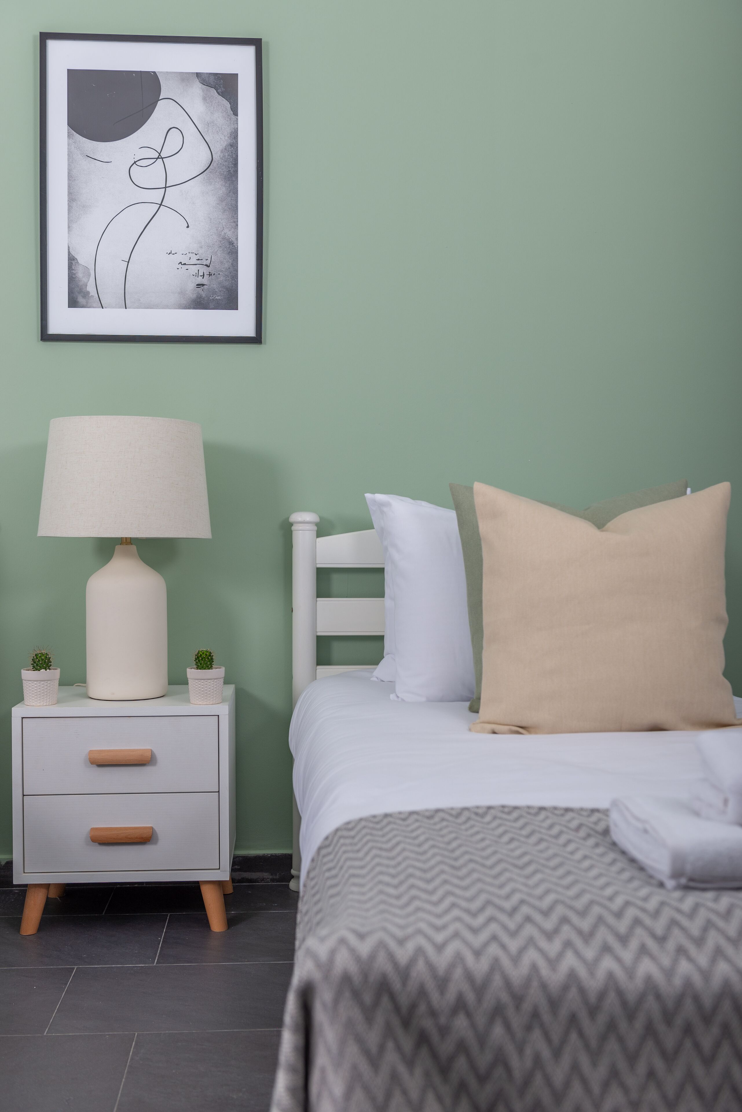 Un dormitorio ordenado con cama, mesita de noche, lámpara y una obra de arte abstracta enmarcada en la pared.