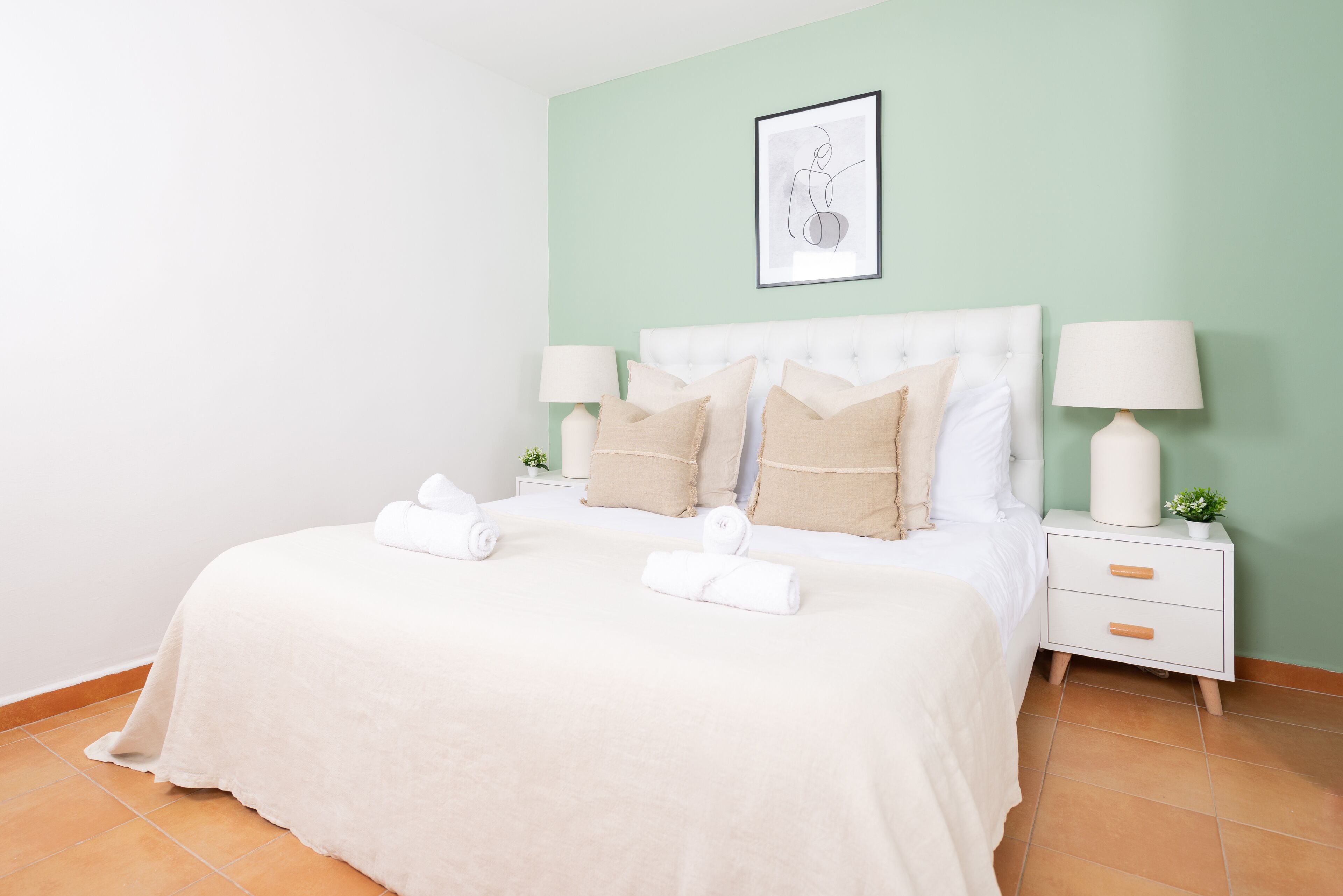 A neatly made bed with white linens and beige pillows, two bedside tables with lamps, and a framed artwork on the wall.