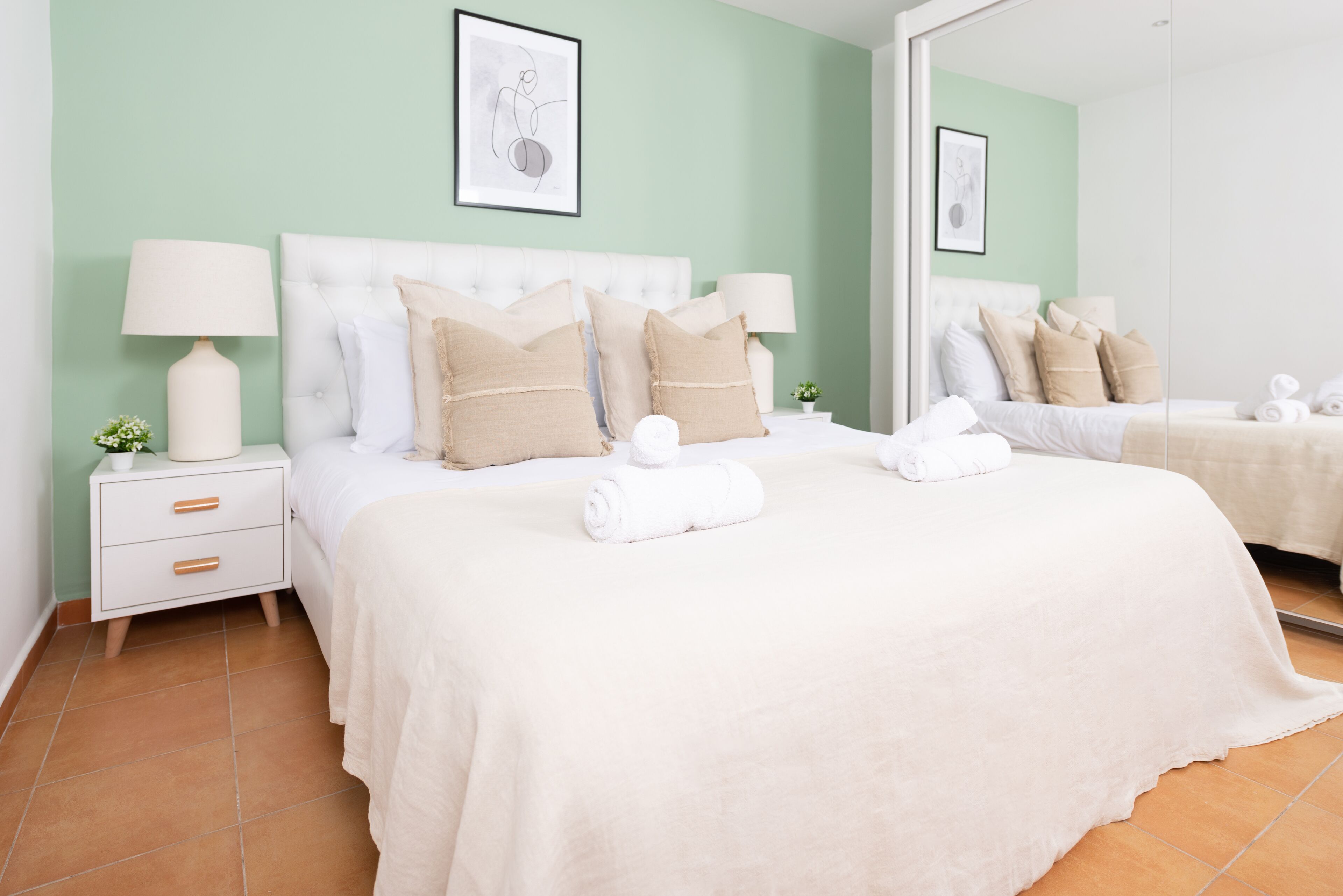 Een net opgemaakt tweepersoonsbed met witte lakens en beige kussens, een nachtkastje met een lamp en een kleine plant.