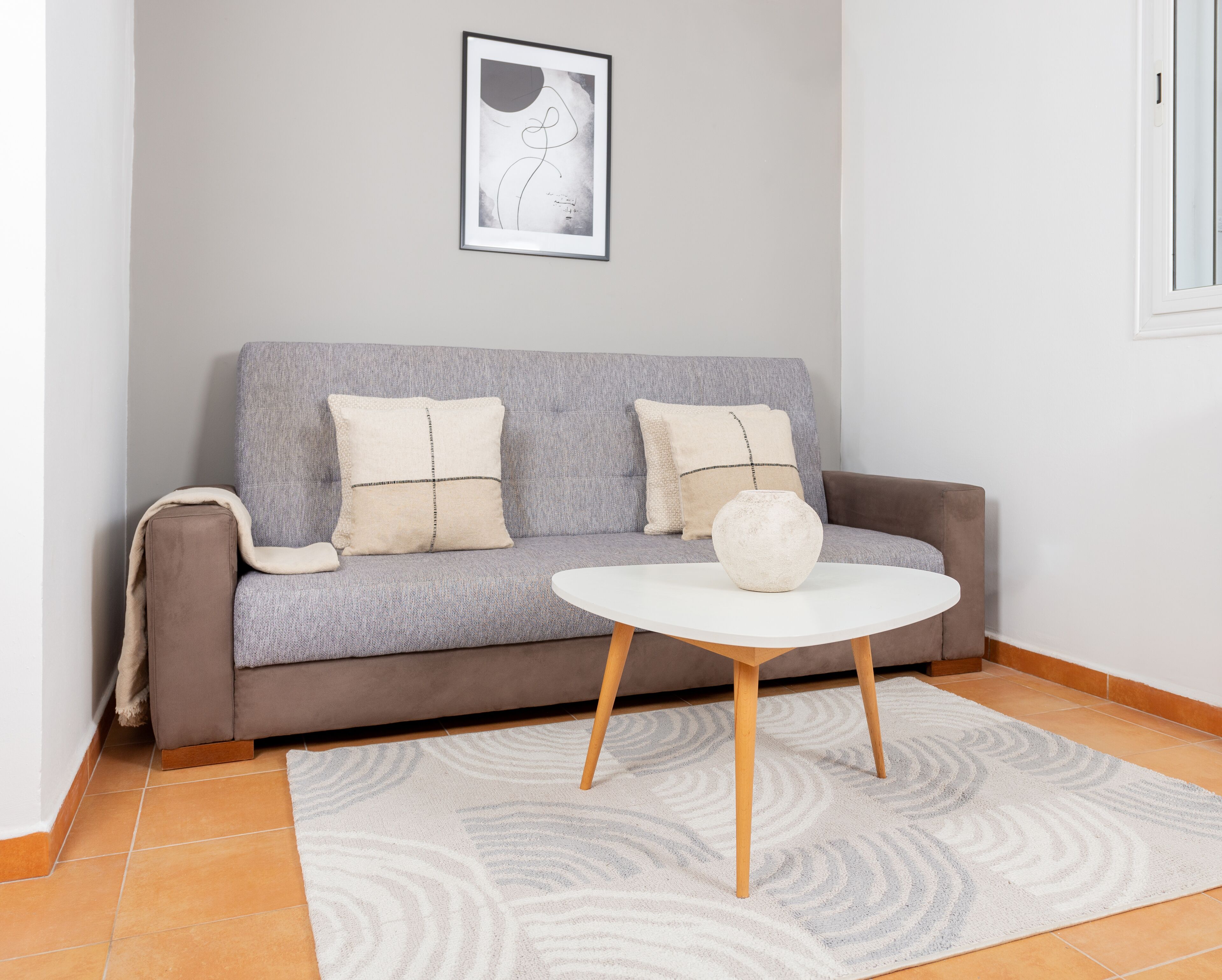 A modern living room with a grey sofa, two white cushions, a white vase, and a round white table.
