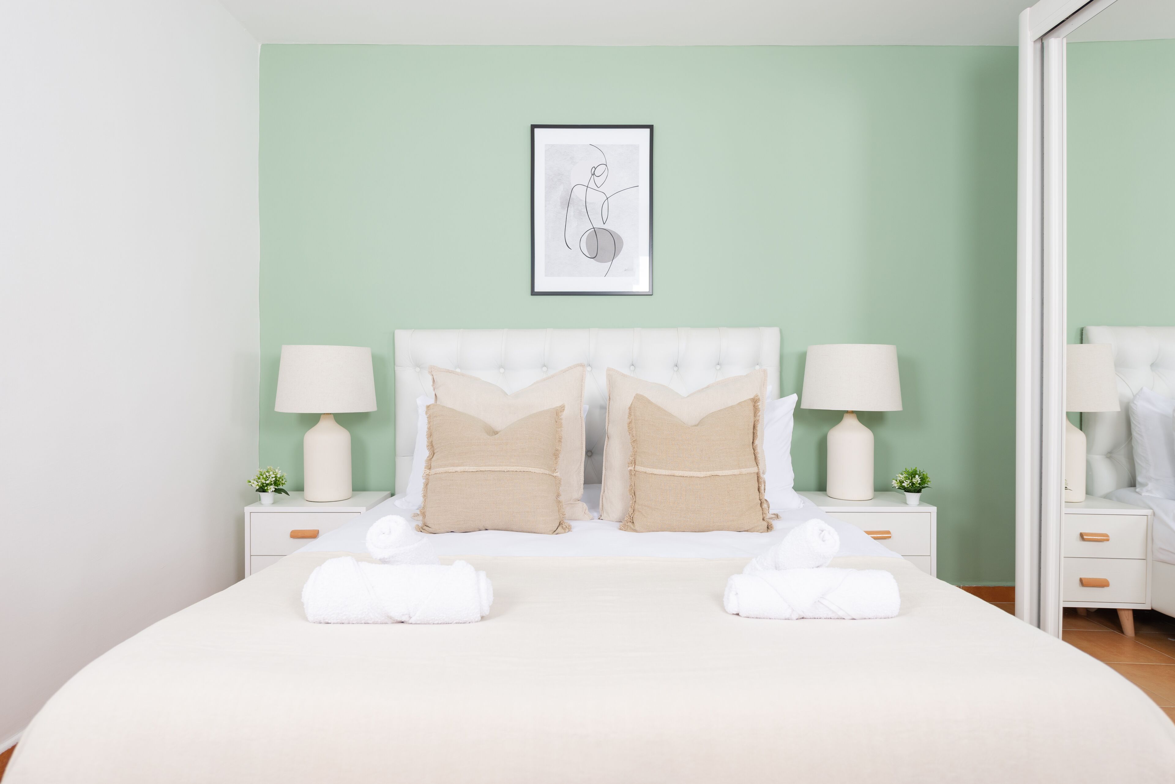 Una cama bien hecha con sábanas blancas y dos almohadas color beige, una obra de arte enmarcada sobre el cabecero y dos mesitas de noche con lámparas y plantas pequeñas.