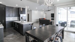 A modern kitchen with dark cabinetry, a central island, and a dining area with a large table and chairs. - Desert Dreams Moab with Home EV Charging (Moab)