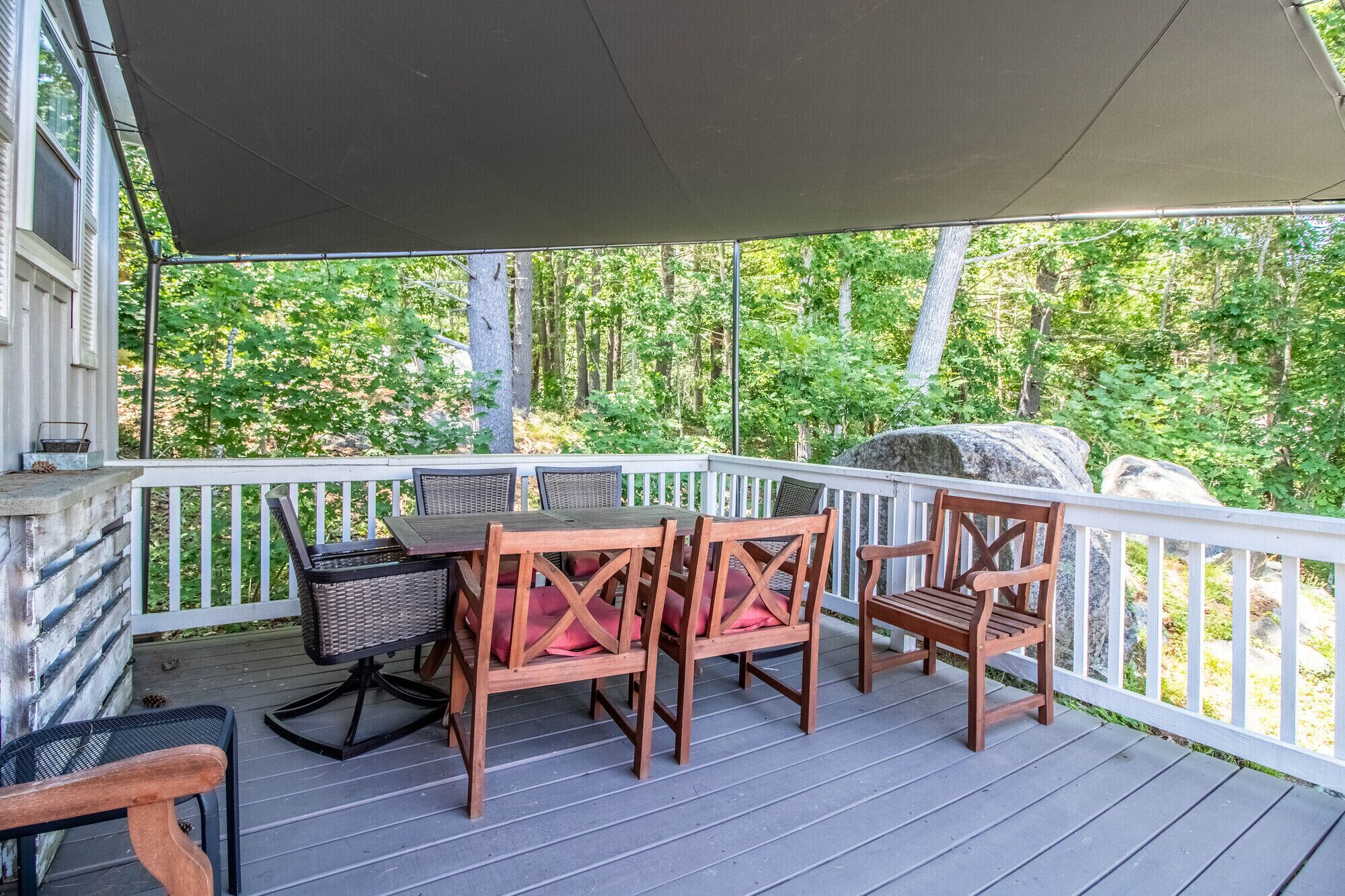Une terrasse extérieure couverte, dotée d’une table et de chaises, entourée d’une balustrade blanche, et offrant une vue sur une zone boisée.