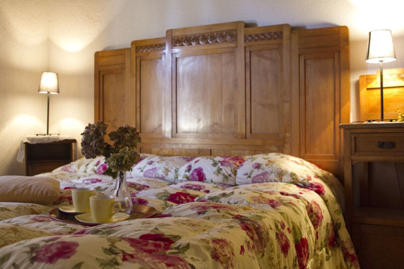 A bedroom with a wooden headboard, floral bedding, a bedside table with a lamp, and a vase with flowers.