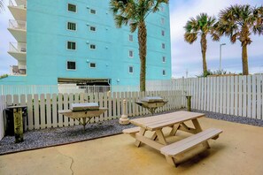 An outdoor patio area with a picnic table and two barbecue grills, surrounded by a white picket fence and palm trees. - Crystal Shores West 507 by Liquid Life (Gulf Shores)