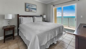 Ein Schlafzimmer mit einem großen Bett, zwei Nachttischlampen, einem Deckenventilator und Blick auf den Ozean durch eine Schiebetür.