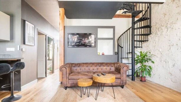 Un salon avec un canapé en cuir marron, deux petites tables d’appoint rondes en bois, un escalier en colimaçon noir et une plante en pot.