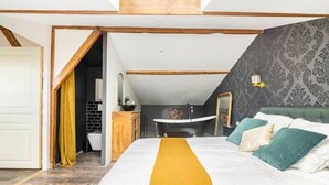 Une chambre avec un grand lit, une tête de lit en bois, une salle de bain avec un lavabo et un miroir, et un mur décoré d’un motif.