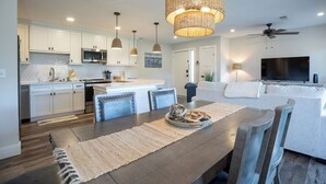A modern kitchen with white cabinets, a dining area with a wooden table, and a living room with a sofa and TV. - 92 Forest Gardens: Newly Remodeled Villa, Walk to Beach, Pool, Free Bikes (Hilton Head Island)