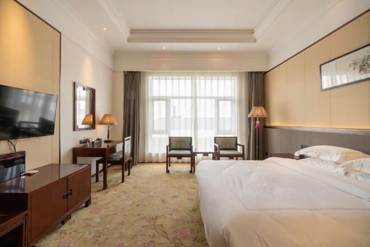 A hotel room with a large bed, a TV, a desk with two chairs, and a window with curtains.