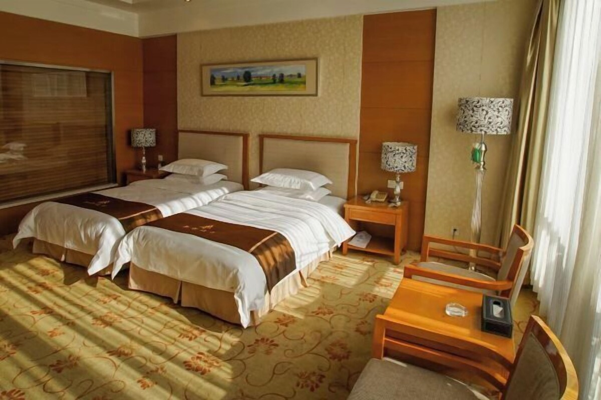 A hotel room with two beds, a desk, a chair, a lamp, and a painting on the wall.