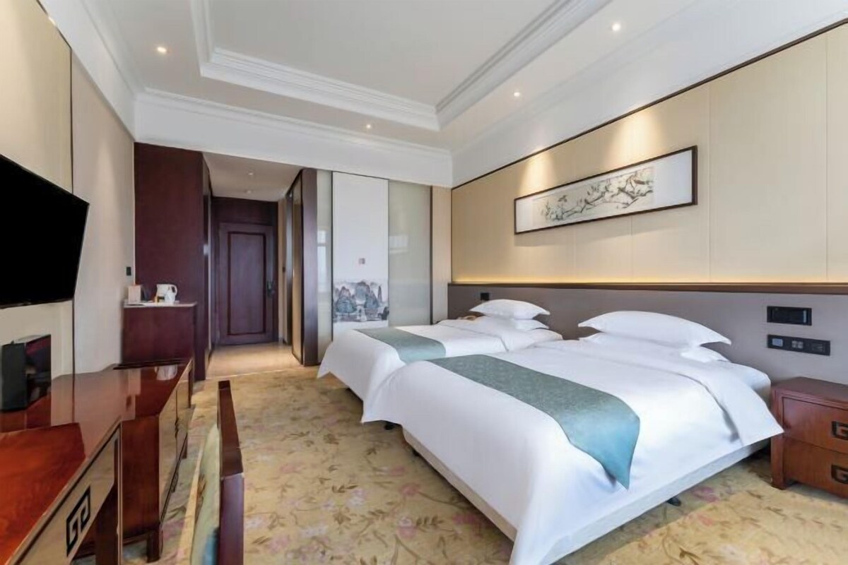A hotel room with two beds, a desk, a television, and a framed artwork on the wall.