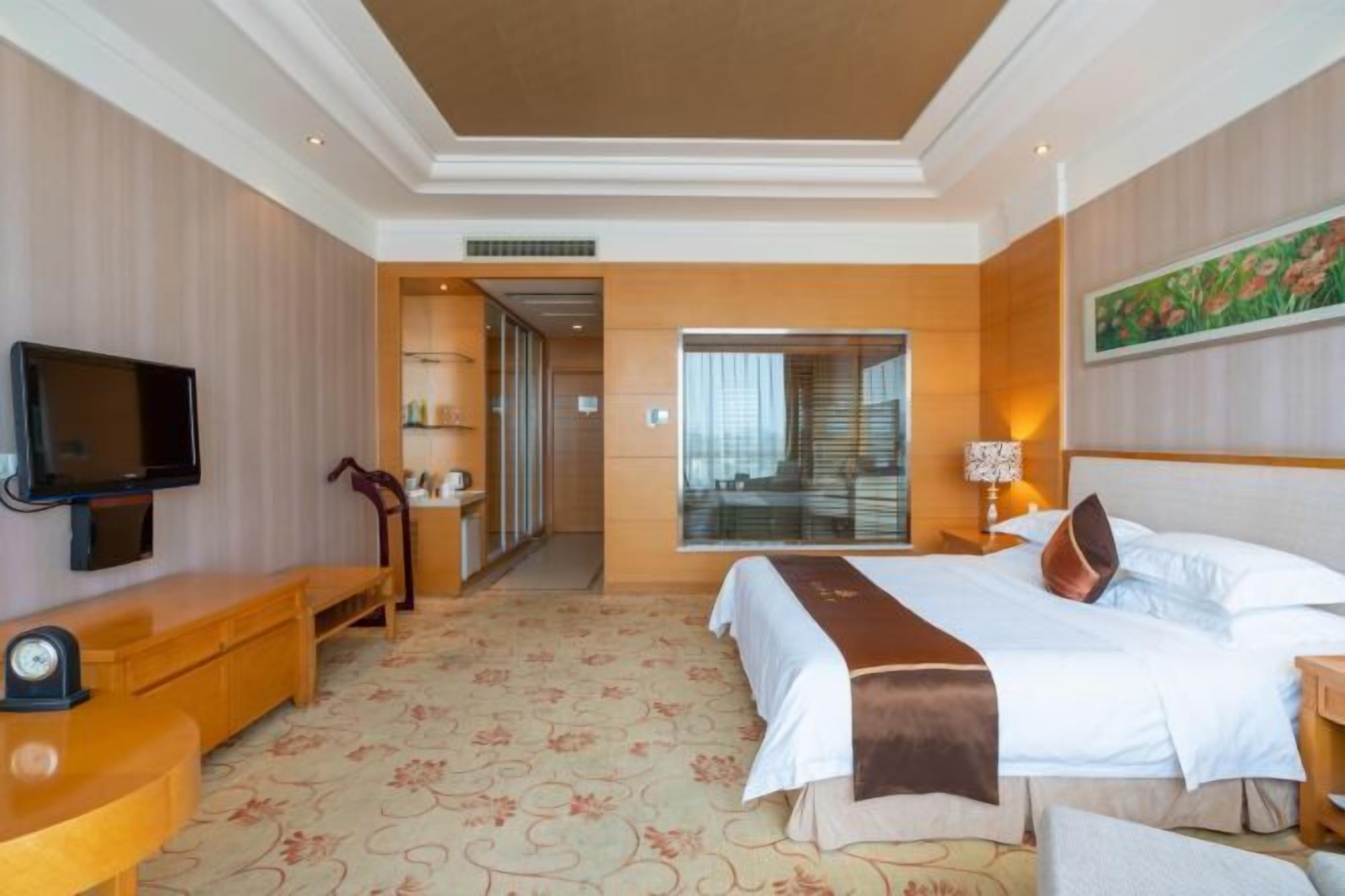 Una habitación de hotel con una cama grande, un televisor sobre un mueble de madera, un escritorio con una silla y un ventanal amplio.