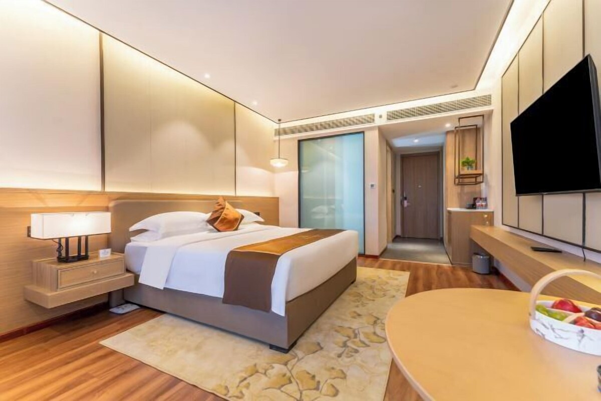 A modern hotel room with a bed, bedside table, lamp, TV, and a fruit basket.