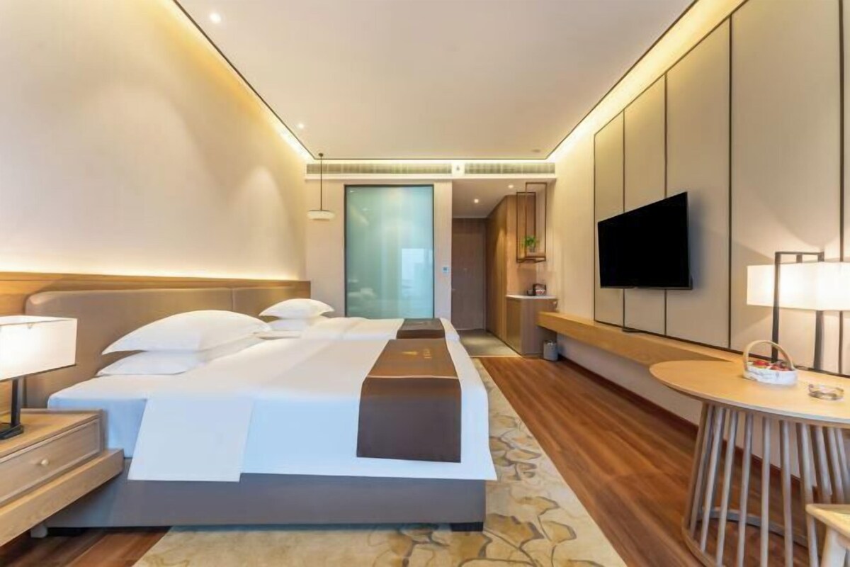 A modern hotel room with a large bed, bedside tables, a TV, and a small round table with a lamp.