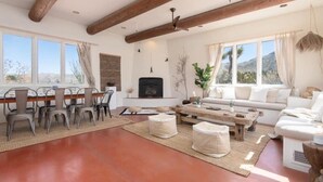 A spacious living room with a fireplace, a sofa set, a dining table with chairs, and large windows offering a view of the outdoors.