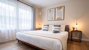 Une chambre à coucher avec un grand lit, deux tables de chevet, une lampe suspendue et une fenêtre ornée de rideaux légers.