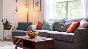 A modern living room with a grey sofa, a blue armchair, a wooden coffee table with a fruit bowl, a floor lamp, and artwork on the wall. - Sanctuary at Oaks | Full Kitchen • Near I75 (Gainesville)
