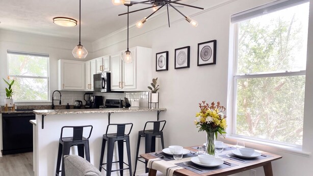A modern kitchen with white cabinets, a central island, bar stools, and a dining table set for two. - Sanctuary at Oaks | Full Kitchen • Near I75 (Gainesville)