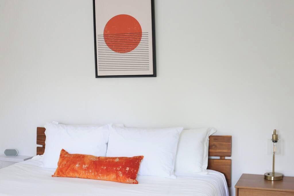 Una cama bien tendida con almohadas blancas y anaranjadas, una obra de arte enmarcada encima y una mesita de noche con una lámpara.