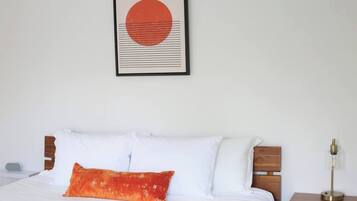 Una cama bien tendida con almohadas blancas y anaranjadas, una obra de arte enmarcada encima y una mesita de noche con una lámpara.