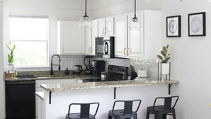 A modern kitchen with white cabinets, a black refrigerator, a built-in oven, and a granite countertop. There are three black bar stools and two pendant lights with exposed bulbs. - Sanctuary at Oaks | Full Kitchen • Near I75 (Gainesville)