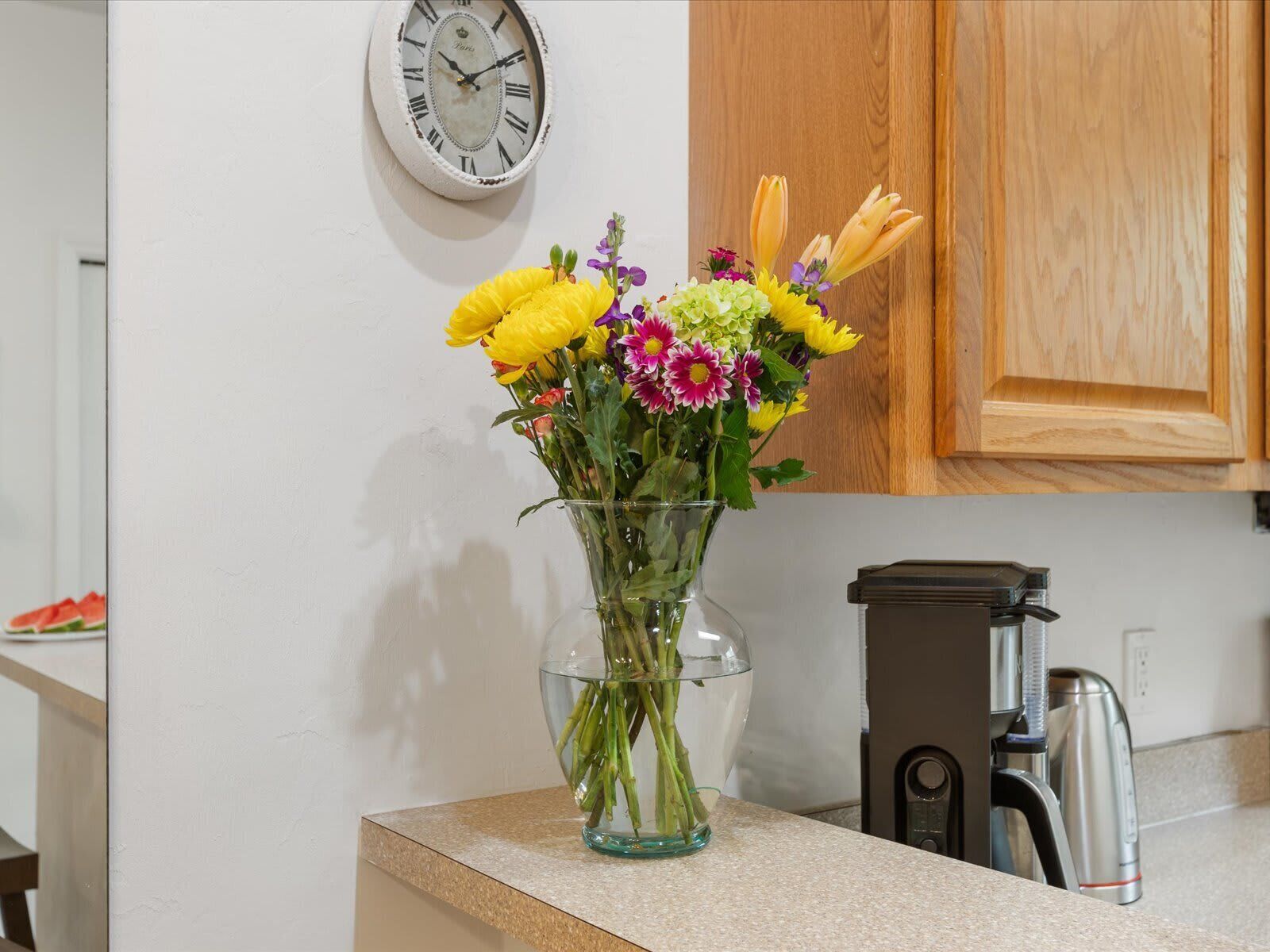 A kitchen with a clock on the wall, a coffee maker, and a vase with flowers on the counter.