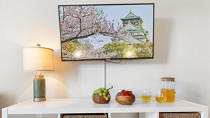 Un televisor montado en la pared muestra un cerezo en flor y un castillo. Debajo del televisor, hay un mueble blanco con una lámpara, un cuenco de uvas, un cuenco de fresas y una jarra de té.