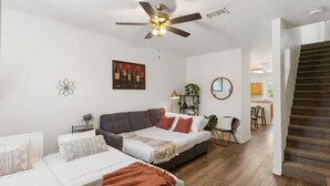 Una sala de estar moderna con un sofá, una mesa de centro y un ventilador de techo. Se ve una escalera que sube a otro nivel.