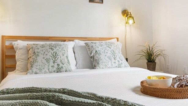 Una cama bien hecha con almohadas estampadas, una lámpara de noche, una planta en maceta y un frutero sobre la mesita de noche.