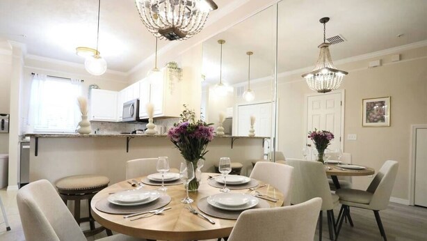 A dining area with a round table set for six, surrounded by six chairs. There are two pendant lights above the table, and a chandelier hanging from the ceiling.