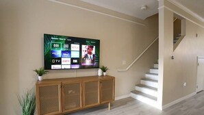 A living room with a mounted TV displaying various streaming services, a wooden console table with plants, and a staircase.