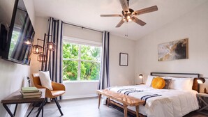 A bedroom with a bed, a chair, a table, a ceiling fan, a window, and a painting.