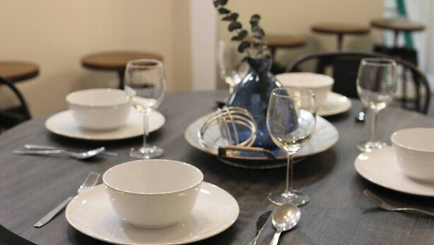 A dining table set with white plates, bowls, cutlery, and wine glasses, with a vase of flowers in the center.