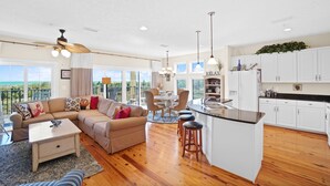 A spacious living area with a sofa, coffee table, and a dining table with chairs. A kitchen with white cabinets and a bar area is visible. - August 2026 Rate Drop! Serendipity by the Sea - 3BR/2BA (Port St. Joe)
