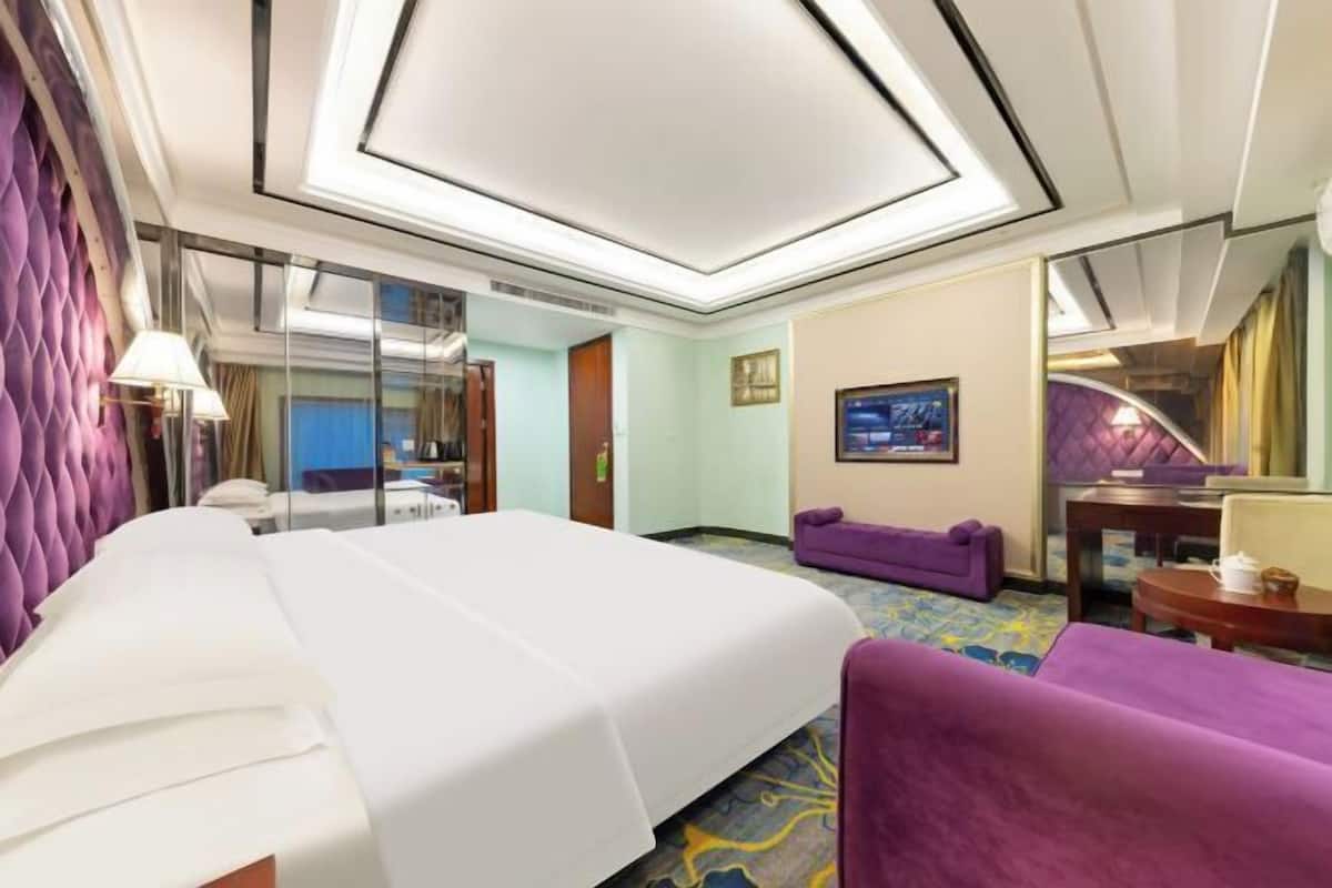 A hotel room with a bed, a purple sofa, a TV, and a mirror.